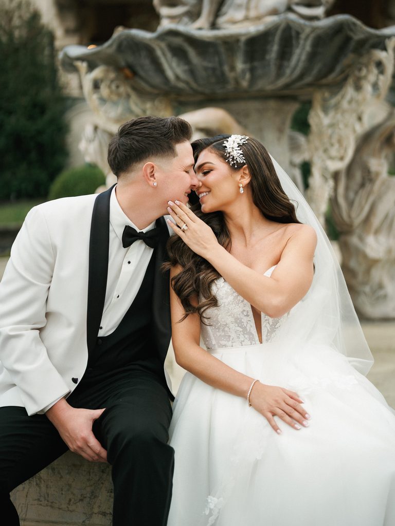 Sophisticated bride touching groom chin near an estate fountain showing her diamond engagement ring and luxury hair piece accessory