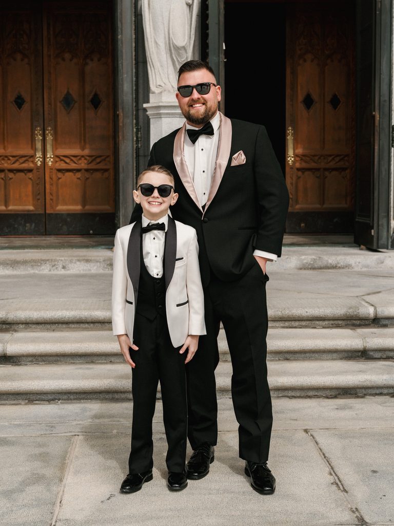 Stylish guest posing with a young ring bearer in matching tuxedos and sunglasses outside a church entrance
