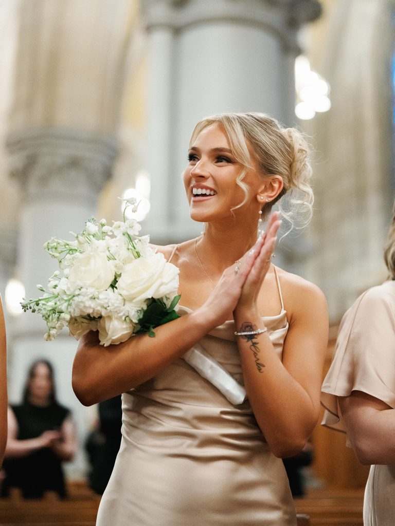 Beautiful blonde bridesmaid in a silk slip dress smiling and clapping with a white rose bouquet inside a grand cathedral
