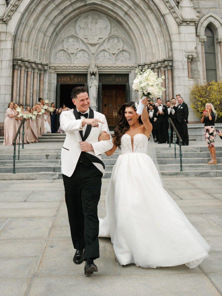 Joyful bride raising her bouquet and groom pointing forward celebrating their wedding exit in front of a historic stone church facade
