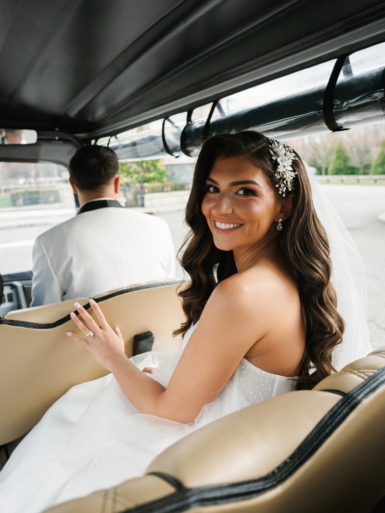 Cinematic luxury bride smiling looking back from a golf cart showcasing her diamond wedding ring and bridal hair piece accessory