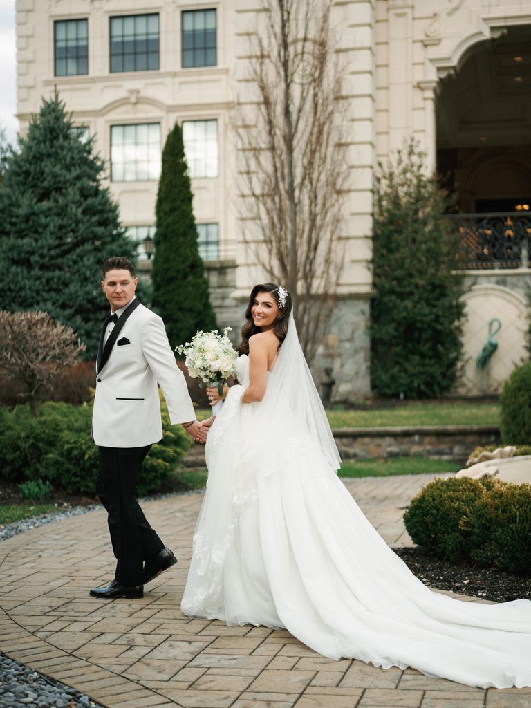 Stunning luxury wedding couple portrait featuring a bride looking back with a dramatic long white gown train outside an upscale country estate manor