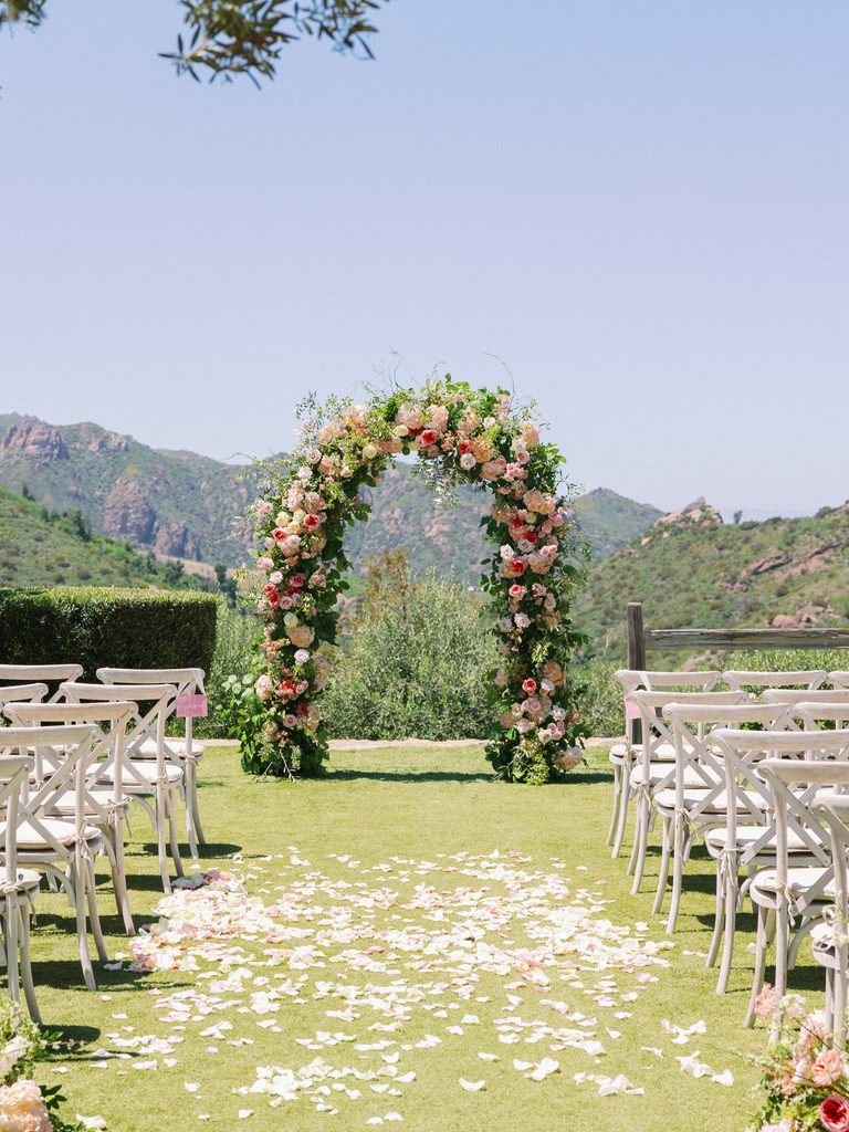 Outdoor wedding altar featuring a lush floral archway and an aisle covered in white petals.