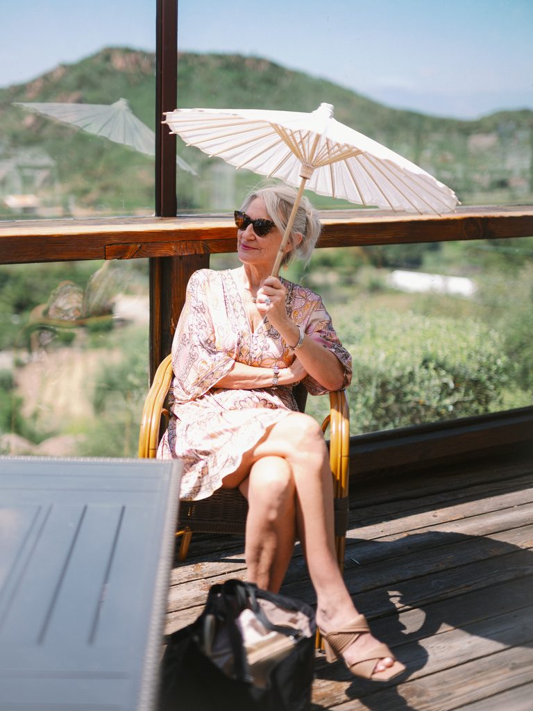Elegant woman in sunglasses sitting on a wooden deck holding a white paper parasol.
