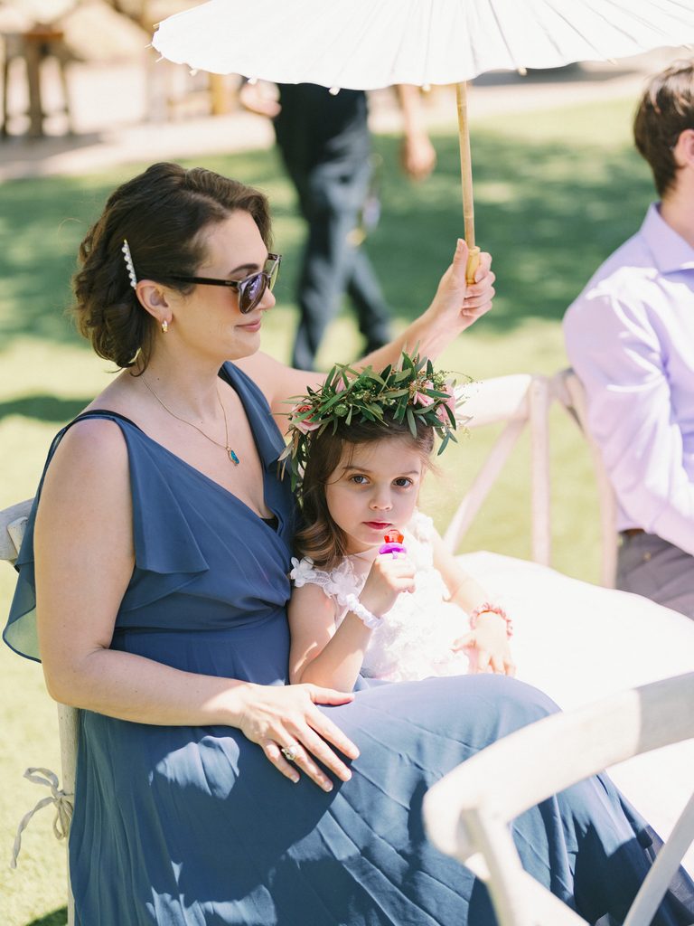 Elegant guest in a blue dress holding a flower girl during a sunny outdoor ceremony.
