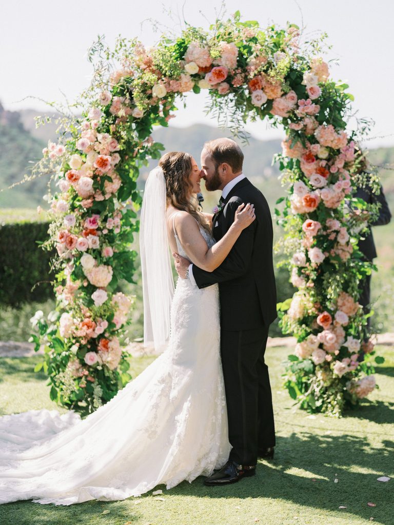 Newlyweds kissing under a massive pink and peach floral arch, a signature shot in a wedding photo gallery.