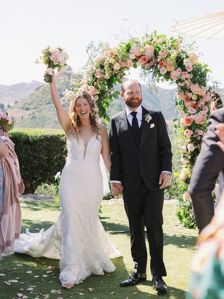 Joyful bride raising her bouquet while walking down the aisle with the groom.