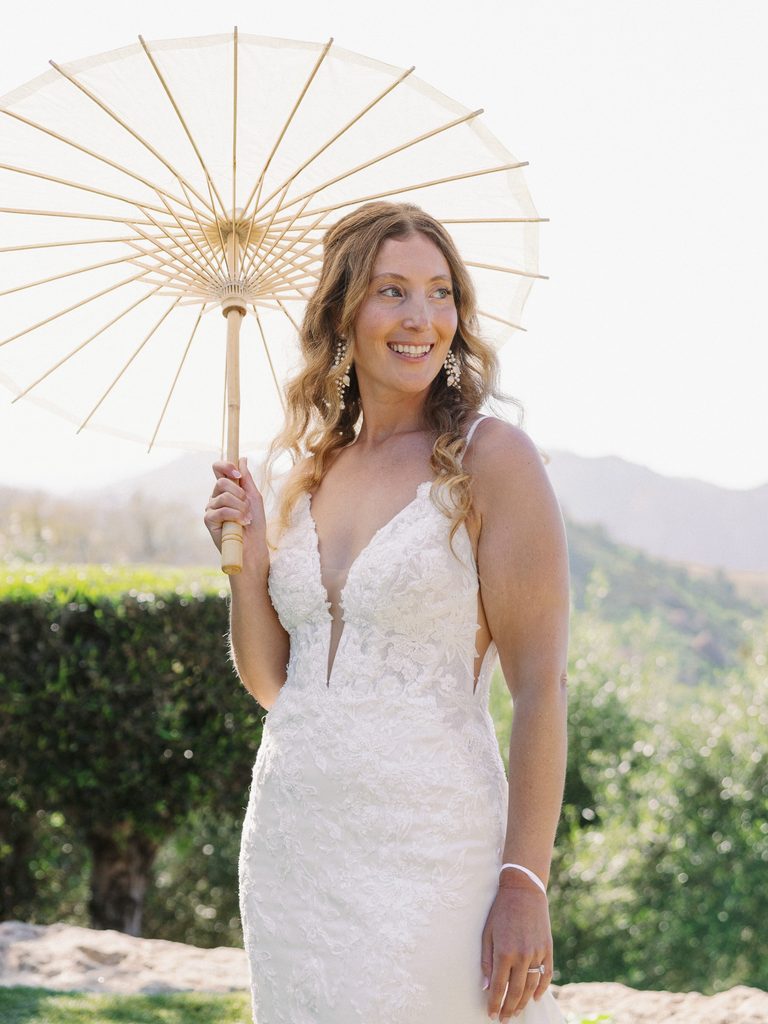 Smiling bride holding a white paper parasol against a soft mountain backdrop.