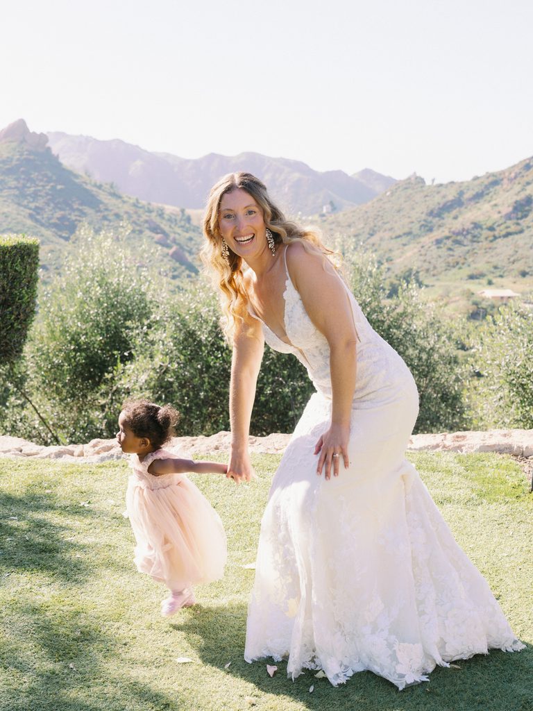 Laughing bride holding hands with a little flower girl on a sunny lawn.