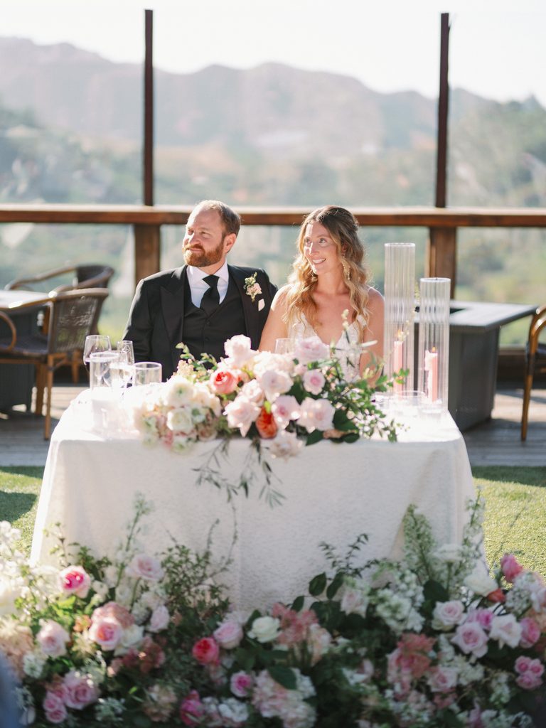 Bride and groom sitting at a decorated sweetheart table with pink florals during an outdoor reception.