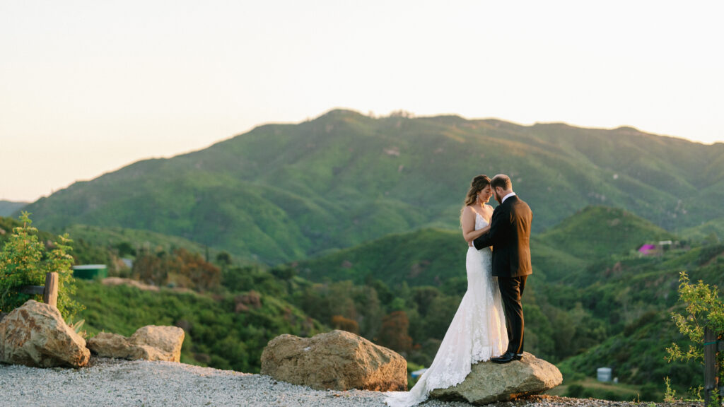 Bride and groom standing on a rock overlooking rolling green mountains at golden hour.