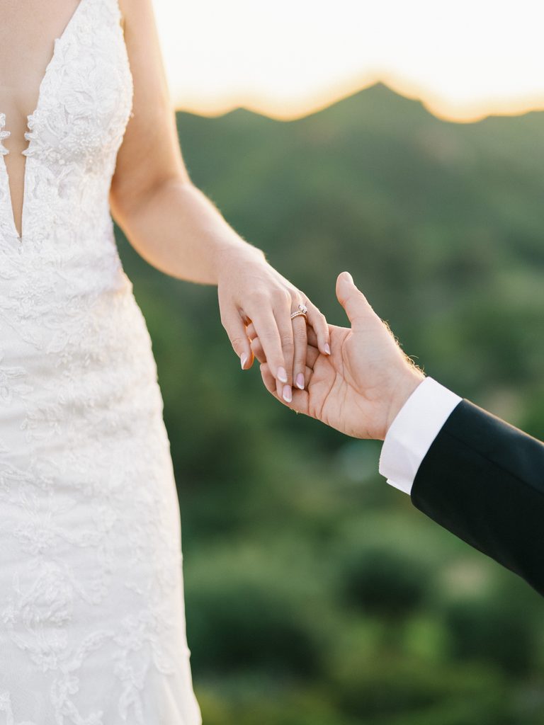 Close-up of a bride and groom holding hands, highlighting the diamond engagement ring during golden hour.