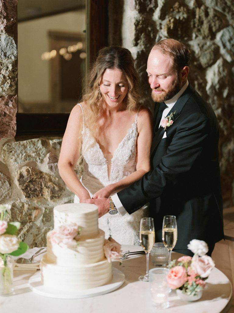 Newlyweds smiling together while cutting their white tiered wedding cake.