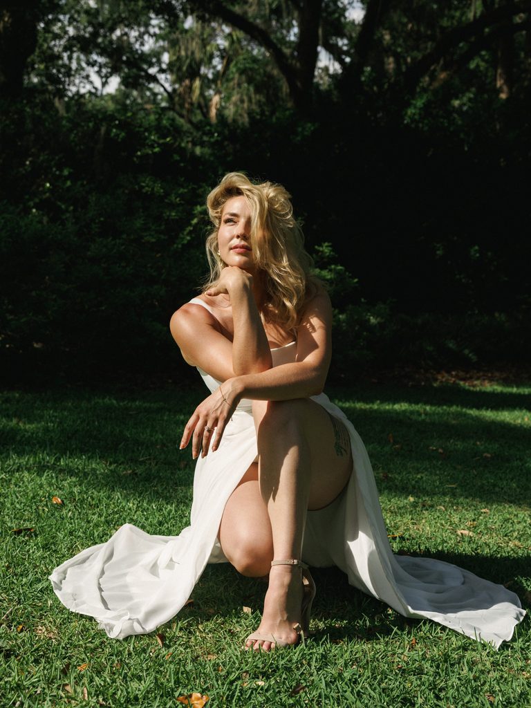 Full-length portrait of a blonde bride-to-be posing in a long white dress on a lawn with trees behind.