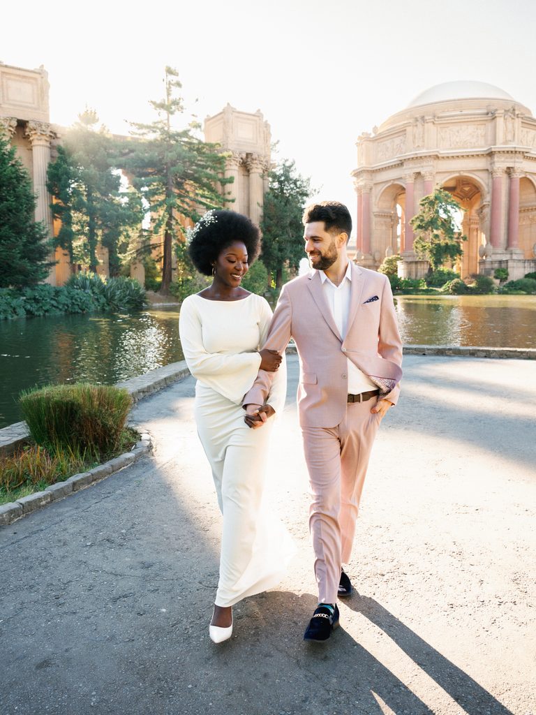 Bride-to-be in a white long-sleeve dress and groom-to-be in a dusty pink suit walking near a pond, showcasing stylish pastel engagement photo outfits.