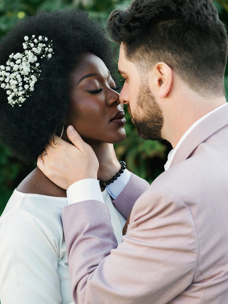 Groom-to-be gently cradling the face of his bride-to-be with closed eyes, highlighting baby's breath flowers in her afro hair.