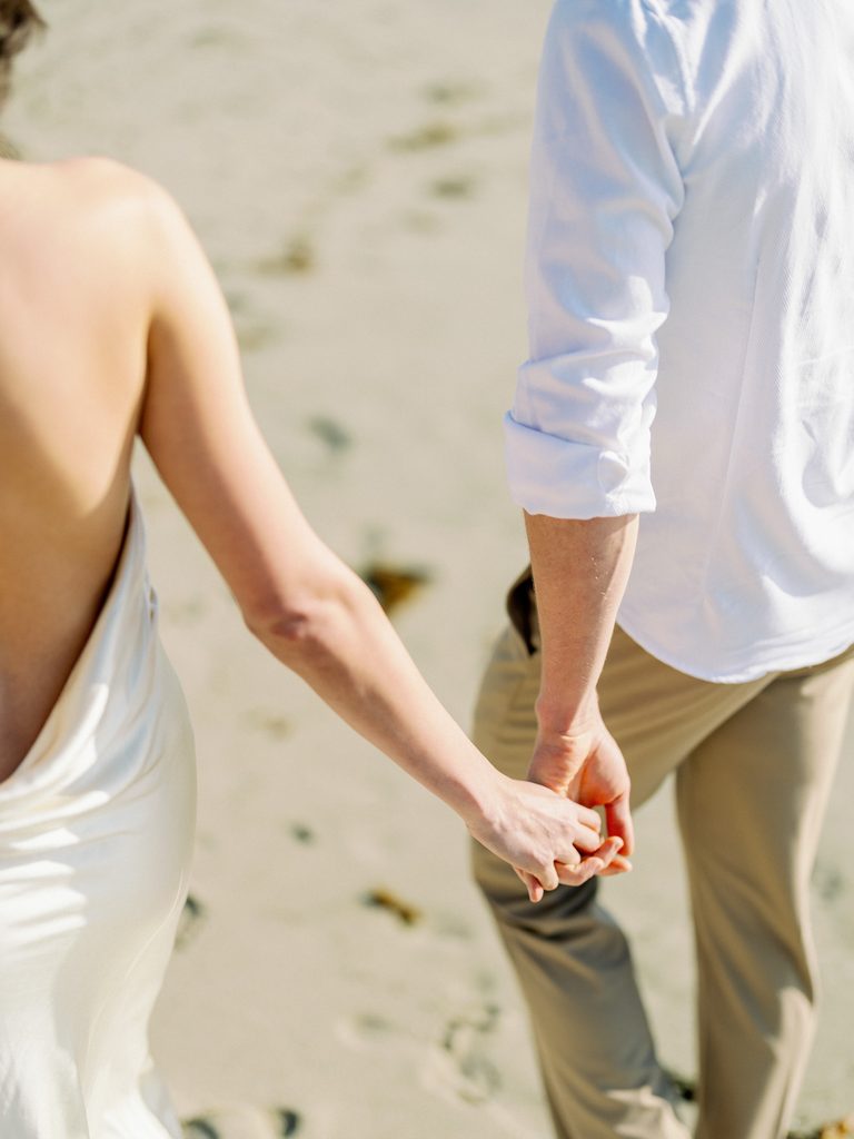 Close-up from behind of a couple holding hands while walking along the shore.