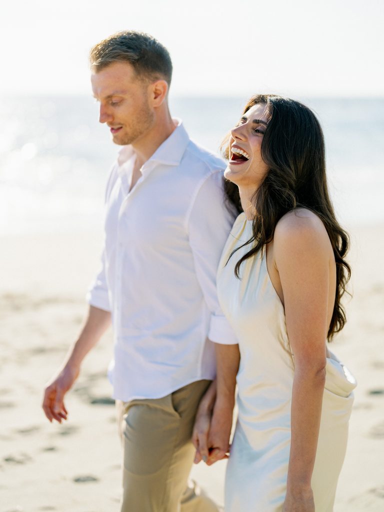 Laughing couple walking hand-in-hand on the beach in effortless summer style.