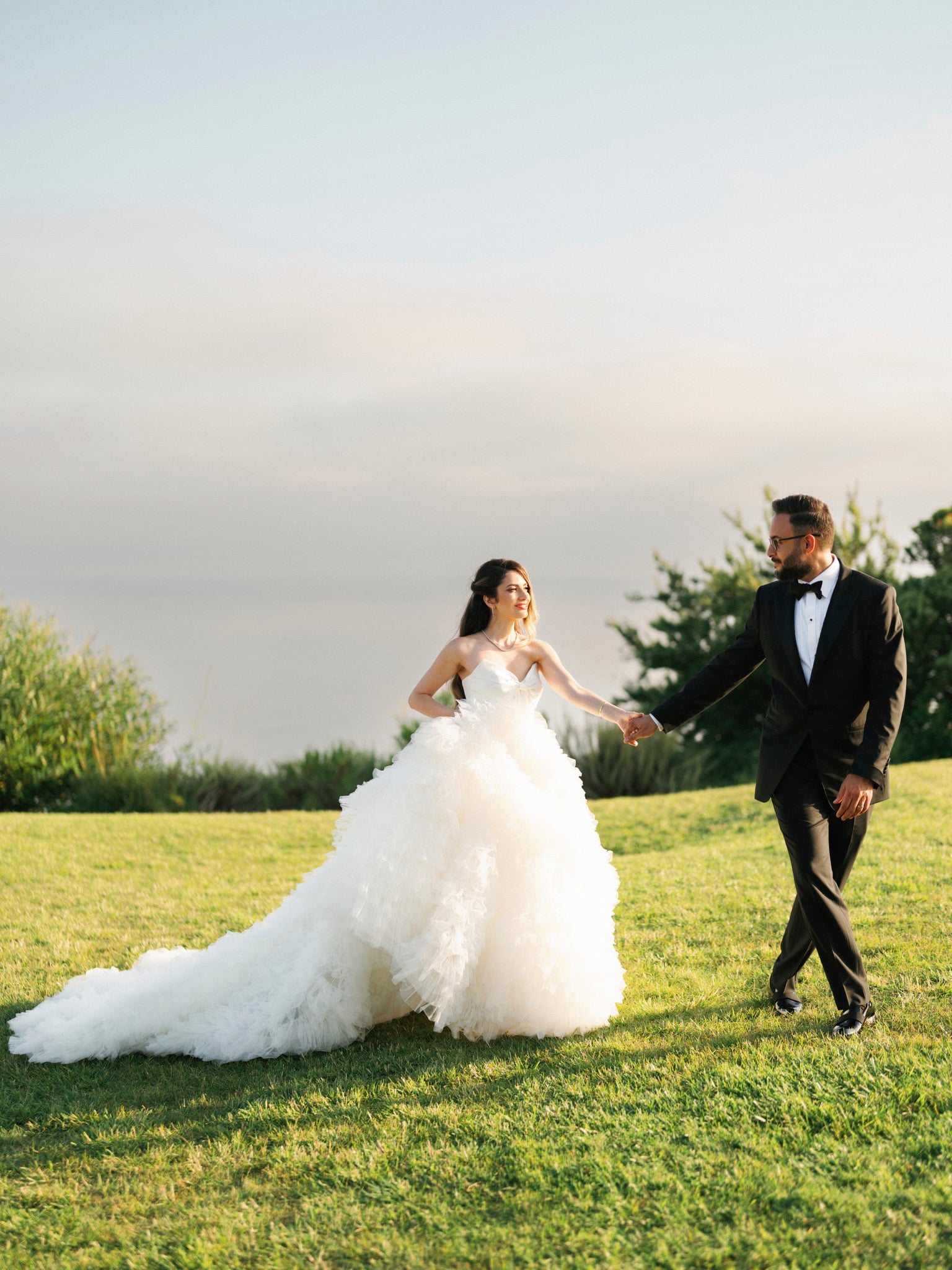 Couple walking hand in hand across a sunlit lawn.