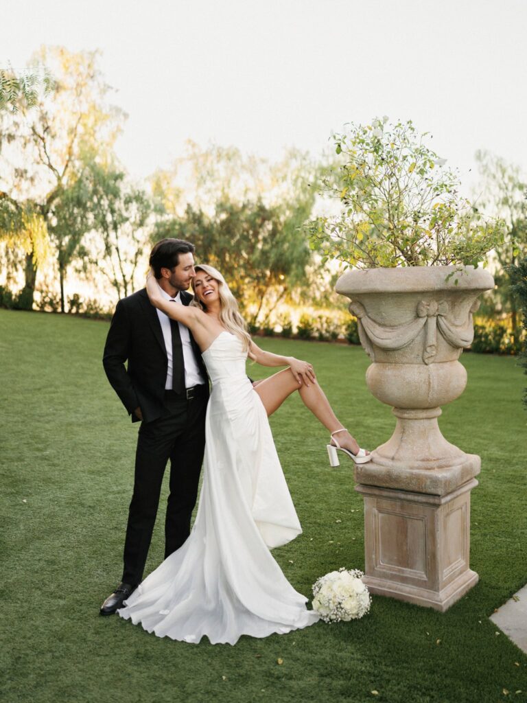 Newlyweds laughing beside a stone garden urn.