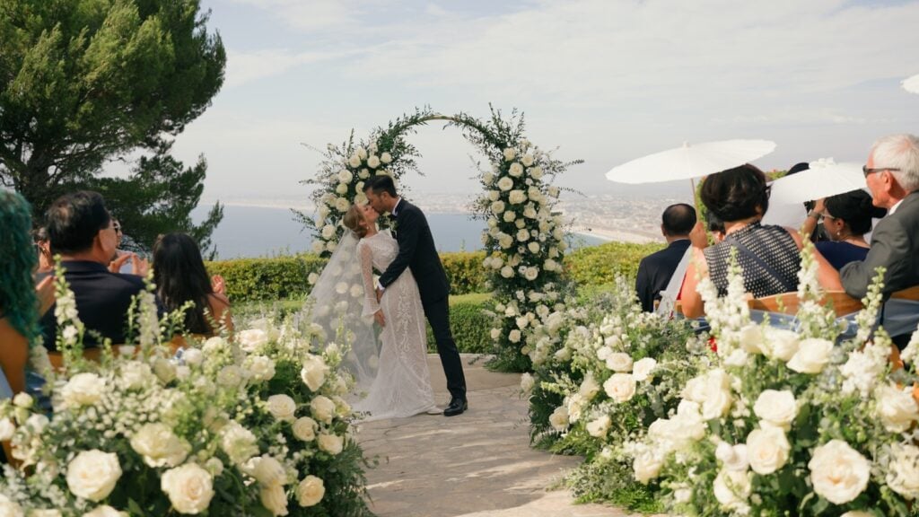 An outdoor wedding ceremony at La Venta Inn, with a white floral arch and guests watching the couple’s first kiss against a scenic coastal backdrop.