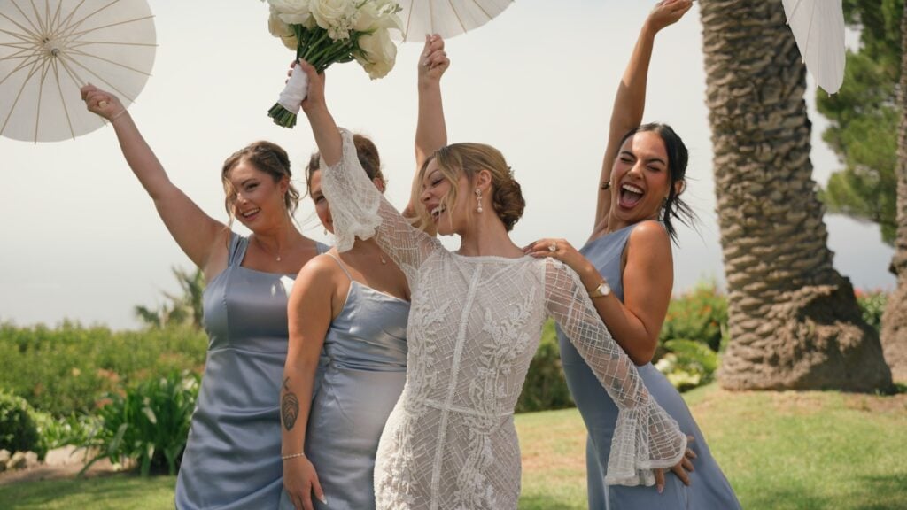 A bride and her bridesmaids in light blue dresses raise their arms and laugh together during a joyful celebration at La Venta Inn, Palos Verdes Estates.