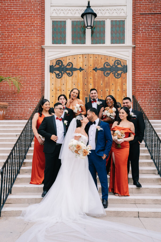 Alondra and Johan sharing a kiss on the steps of a brick building, surrounded by their wedding party in vibrant orange and black attire. A joyful California wedding celebration planned by Not Jess A Planner and captured by Dreamwood Films.