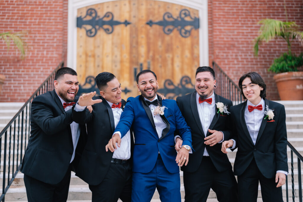 A group of groomsmen in black suits and red bow ties laughing together on a staircase. A fun, candid portrait that reflects the joyful atmosphere of this California wedding celebration, captured by Dreamwood Photo and planned by Not Jess A Planner.