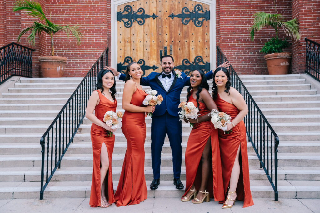 The groom smiling brightly as he poses with the bridesmaids in elegant orange gowns on a staircase. A stylish moment captured by Dreamwood Films, highlighting the personalized wedding planning of Jess Valenzuela.