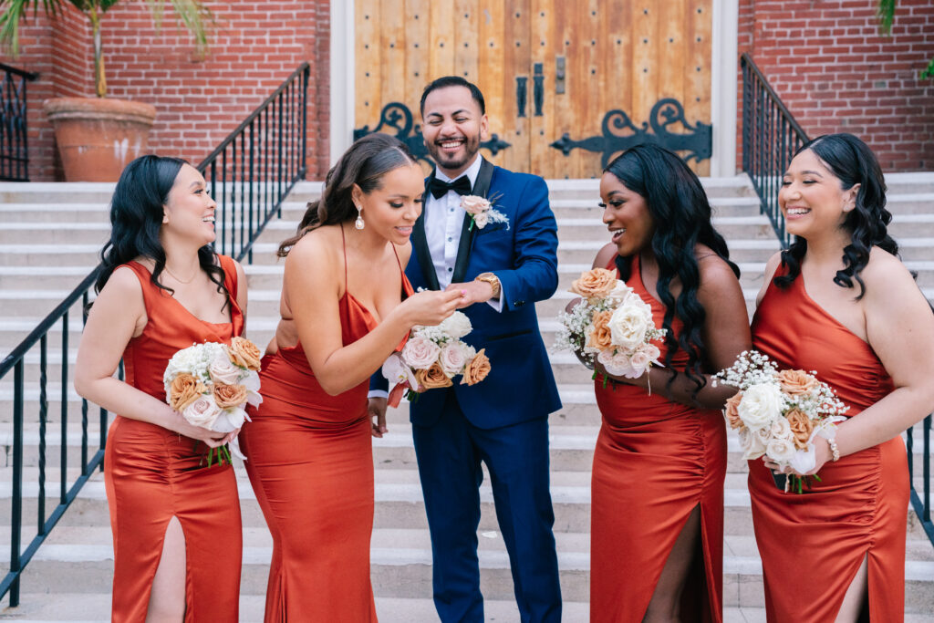 A groom smiling brightly while showing his wedding ring to four bridesmaids holding floral bouquets. A memorable moment from a wedding celebration photographed by Dreamwood Photo.