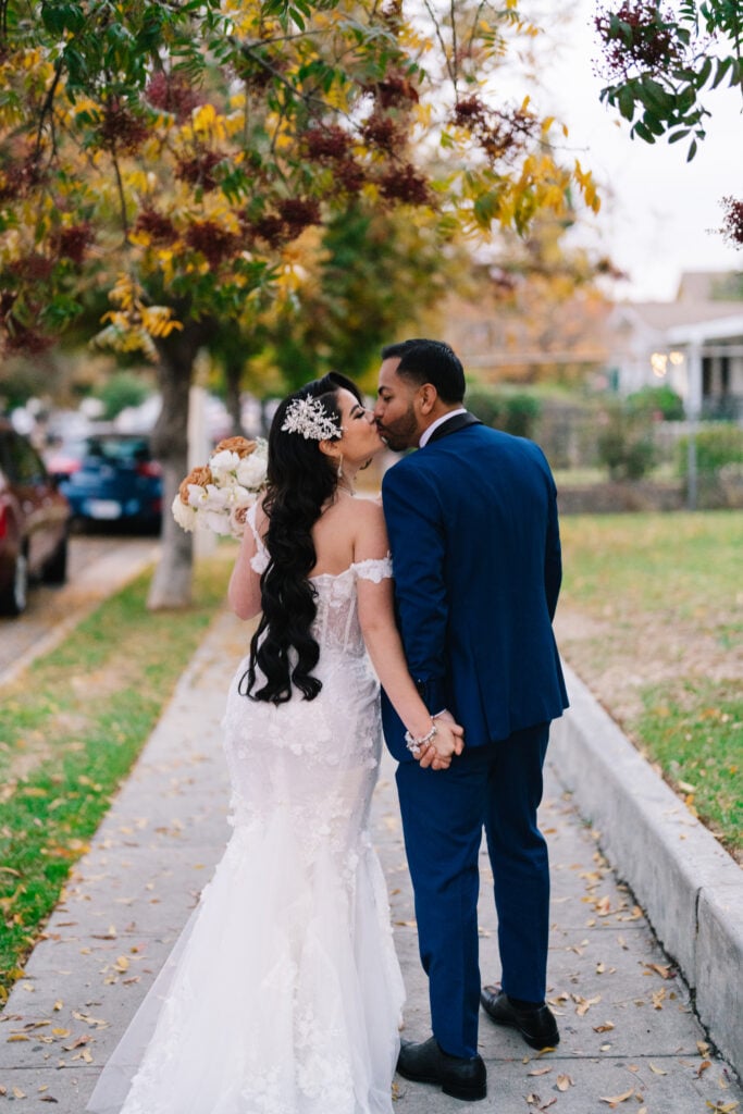 A bride and groom sharing a kiss on a sidewalk beneath autumn trees with yellow and orange leaves. The bride wears a lace mermaid gown and floral headpiece, holding a bouquet while holding hands with the groom in a blue suit. A beautiful California wedding celebration captured by Dreamwood Films and planned by Not Jess A Planner.
