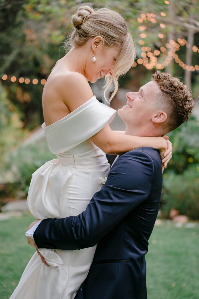 Close up portrait of laughing newlyweds embracing in garden with bokeh string lights