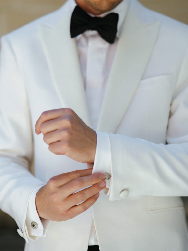 Macro shot of groom hands fixing cuffs wearing luxury white dinner jacket with black velvet bow tie