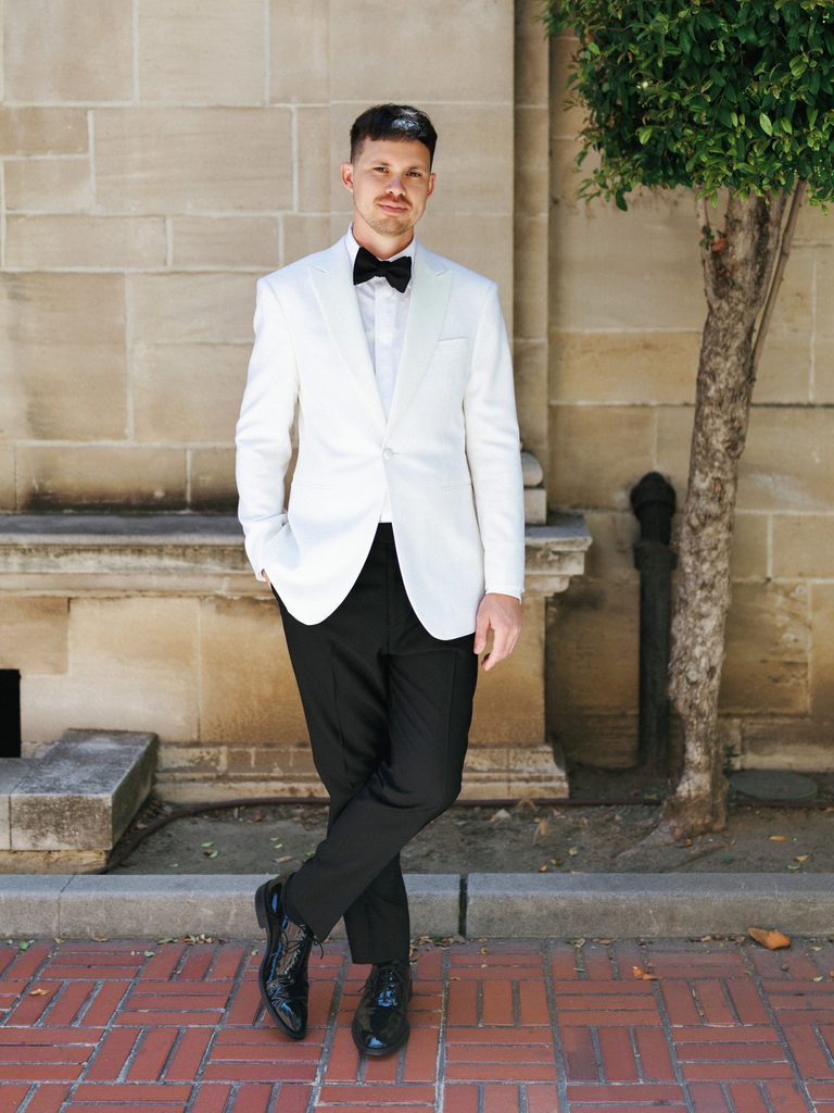 Handsome groom posing in white tuxedo jacket and black trousers outside historic building