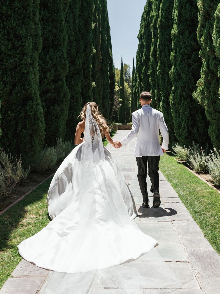 Wide shot of bride and groom running away down stone path between tall green cypress trees