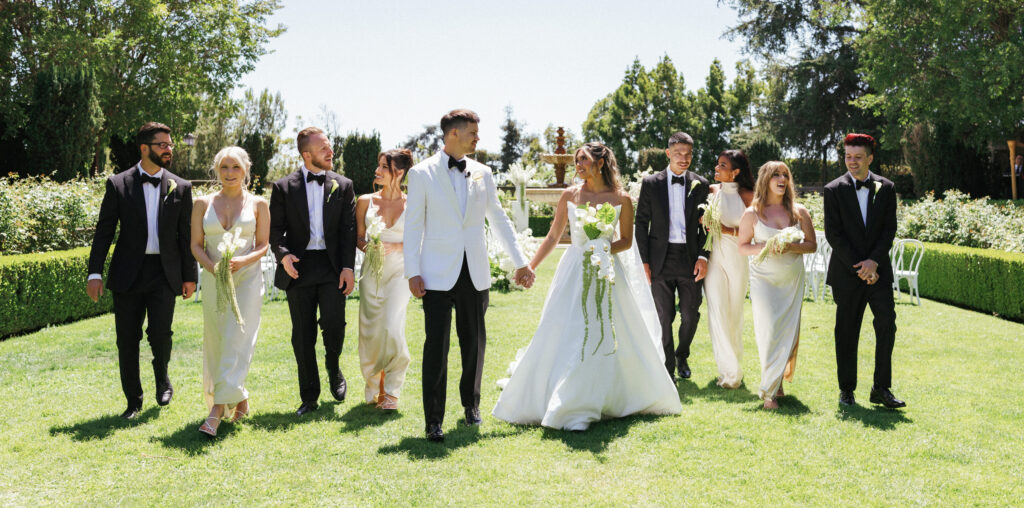 Elegant bridal party walking on green lawn during sunny outdoor wedding photo session