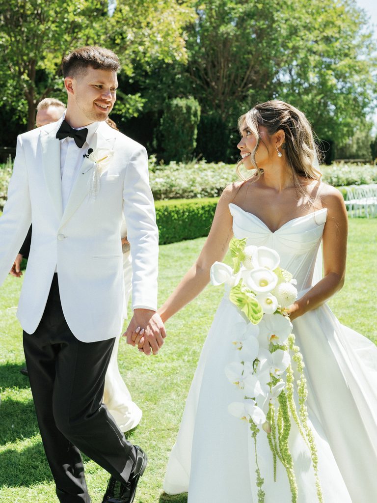 Smiling newlyweds looking at each other outdoors with bride holding cascading white orchid bouquet
