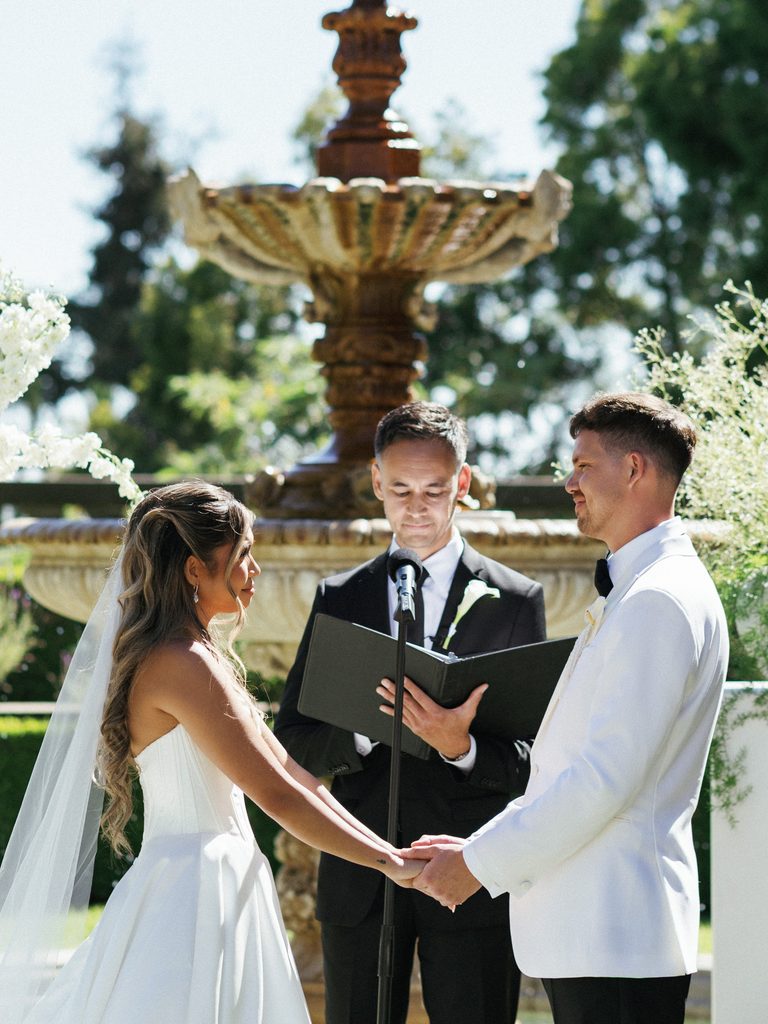 Elegant bride and groom holding hands while listening to wedding officiant during outdoor ceremony