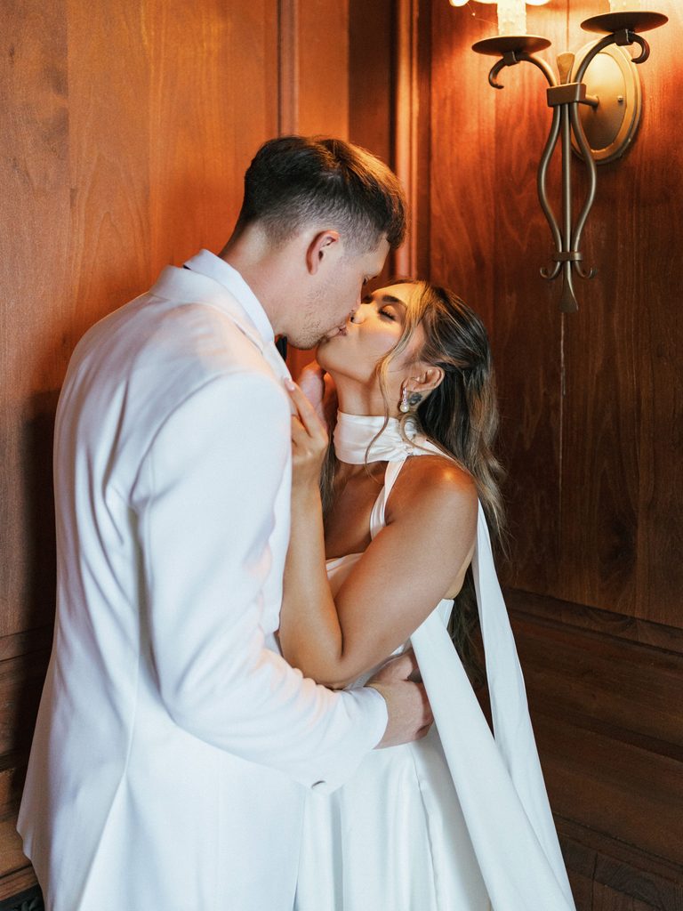 Romantic newlyweds sharing an intimate kiss against dark wood panels with warm wall sconce light