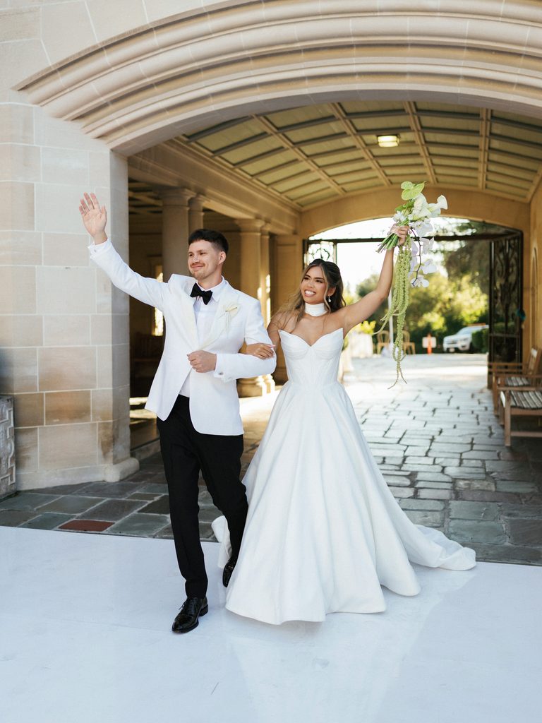 Excited newlyweds waving to guests while entering white dance floor under stone archway