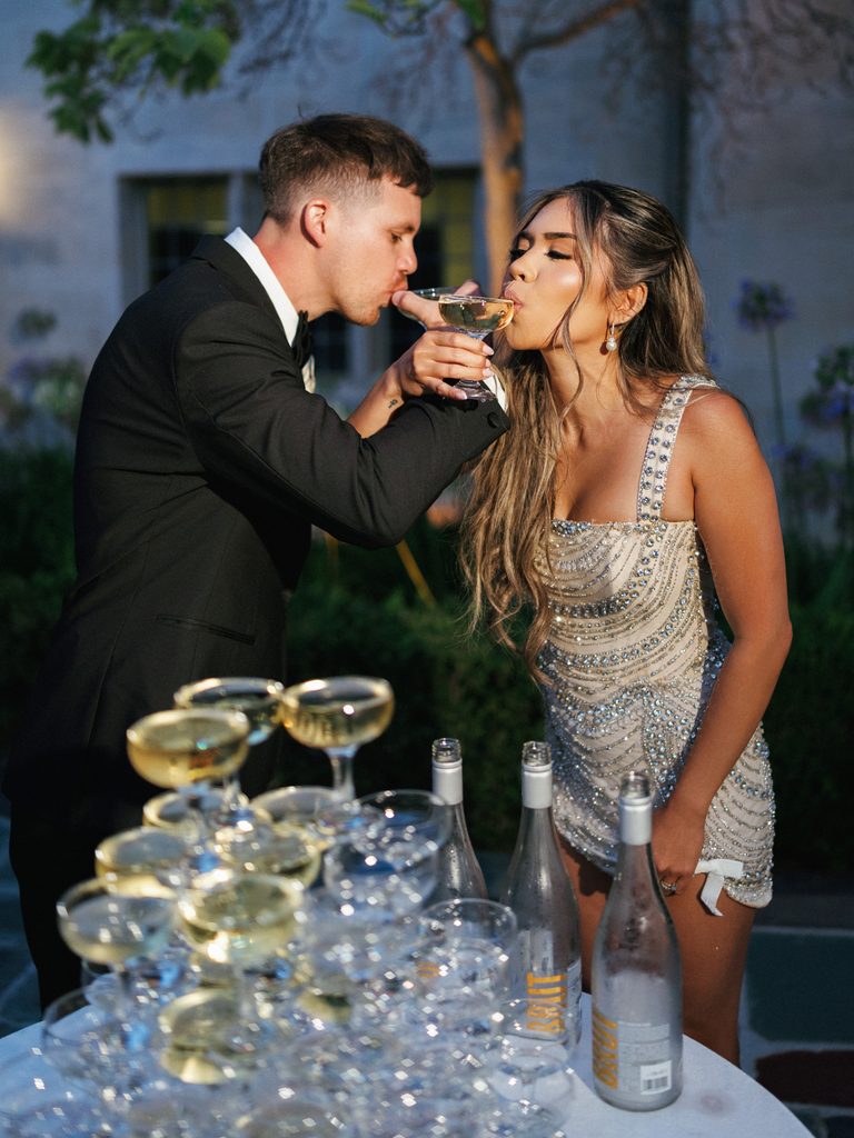 Joyful bride and groom sipping champagne from coupe glasses next to crystal tower at night reception