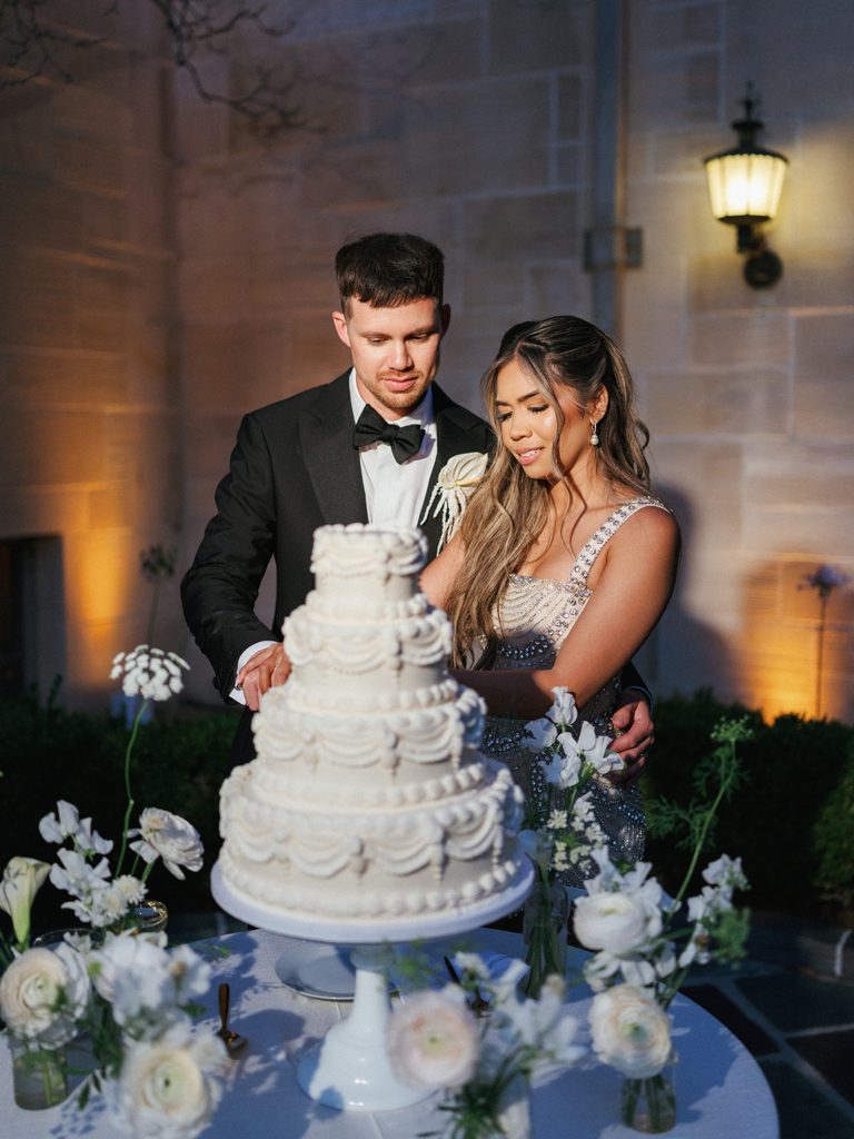 Elegant bride and groom cutting tiered white wedding cake during outdoor night reception