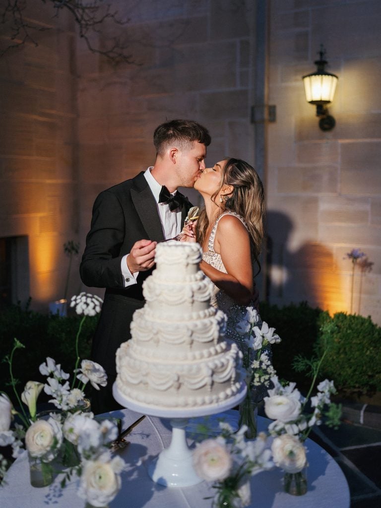 Handsome groom in black tuxedo kissing bride in embellished dress next to luxury wedding cake