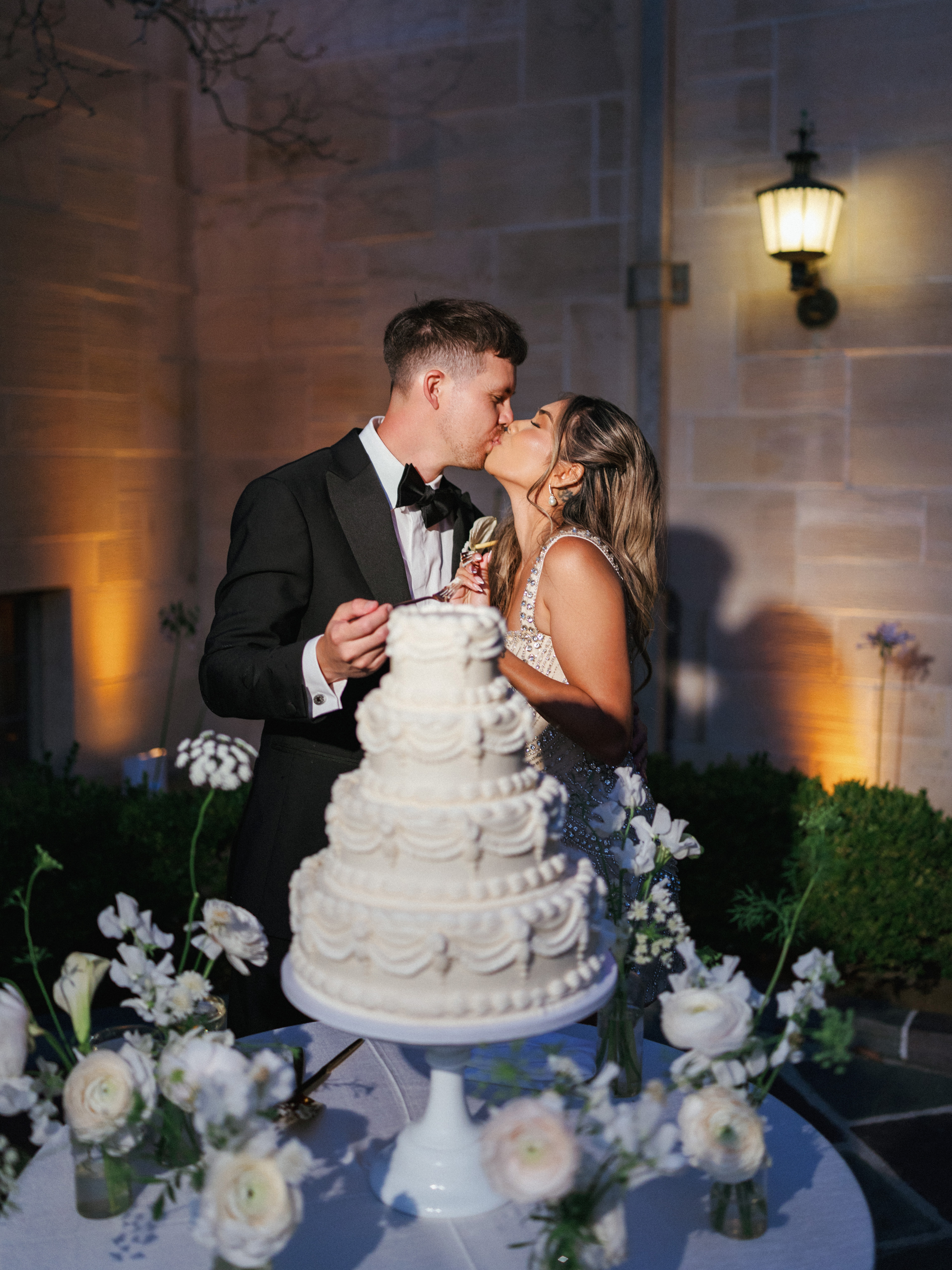 Handsome groom in black tuxedo kissing bride in embellished dress next to luxury wedding cake