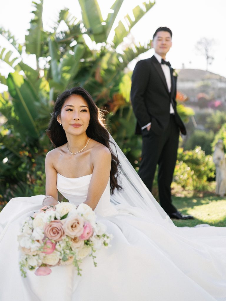 Beautiful asian bride sitting on green lawn in full length white wedding gown with groom standing in background