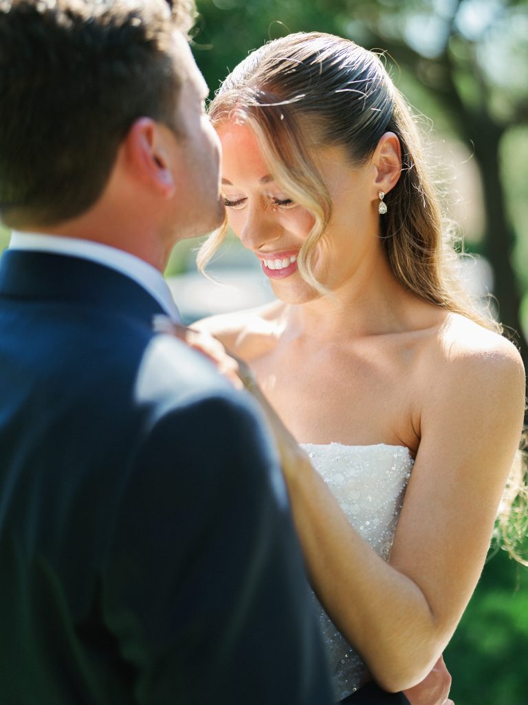 Groom kissing smiling brides forehead during romantic outdoor vineyard wedding photoshoot