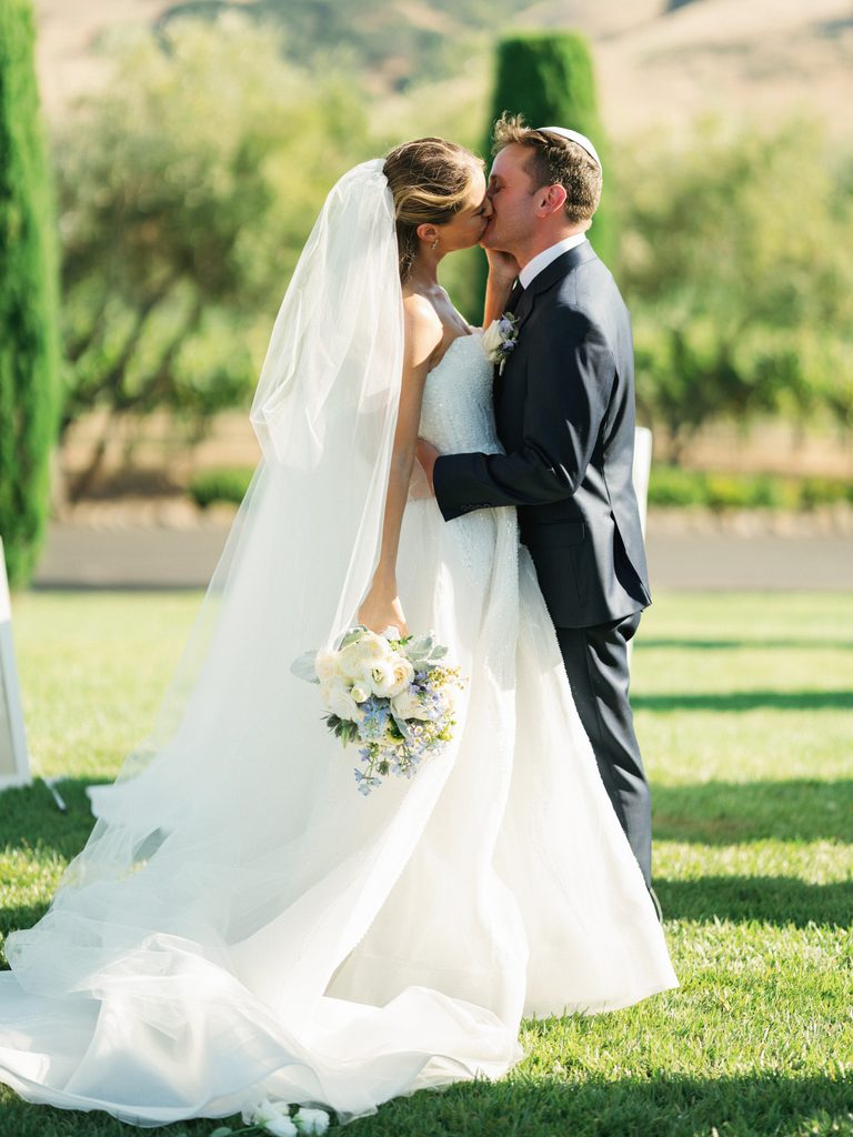 Romantic newlyweds kissing on green grass during luxury outdoor wedding ceremony