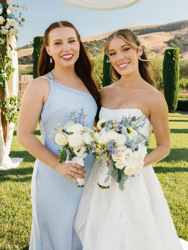 Beautiful bride posing with maid of honor in light blue dress at vineyard venue