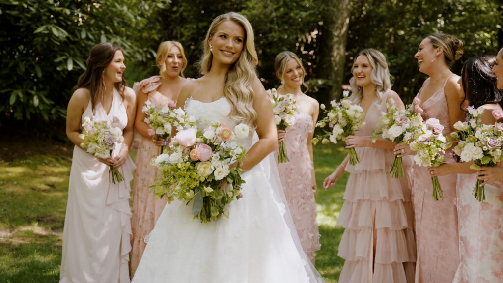 A bride in a white lace gown holding a colorful bouquet, standing with her bridesmaids in blush pink dresses in a sunlit garden.