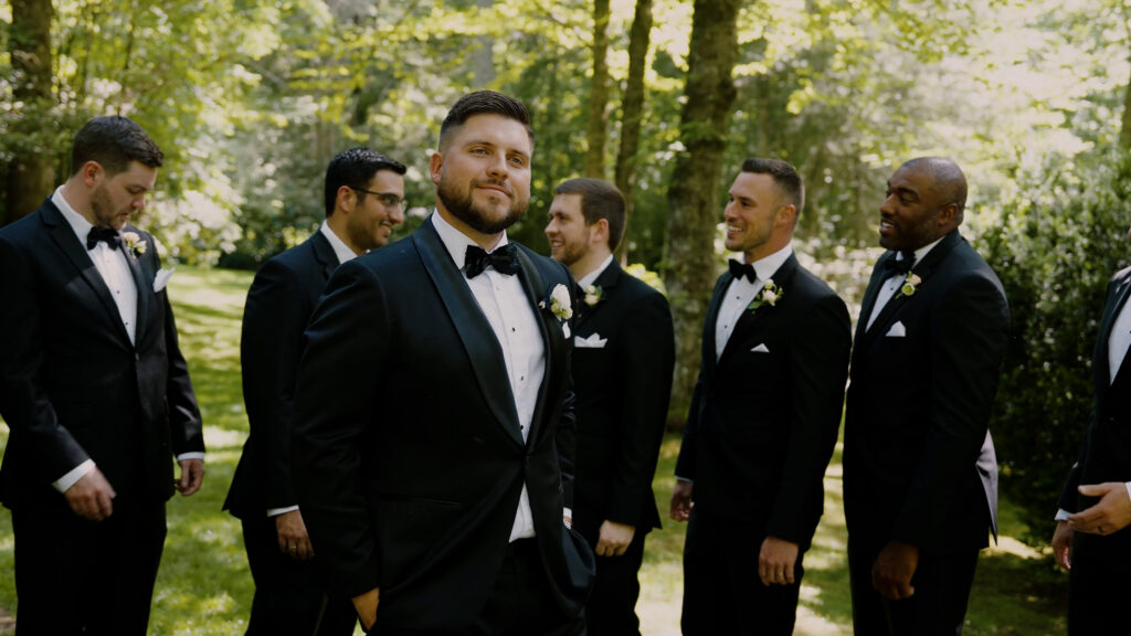A groom in a black tuxedo smiling confidently, surrounded by his groomsmen in matching black tuxedos and bow ties outdoors near trees.